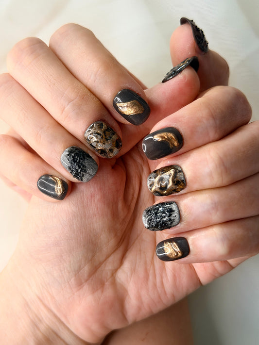 Close-up of hands showcasing a black and gold marble-patterned nail art design with metallic gold foil accents.