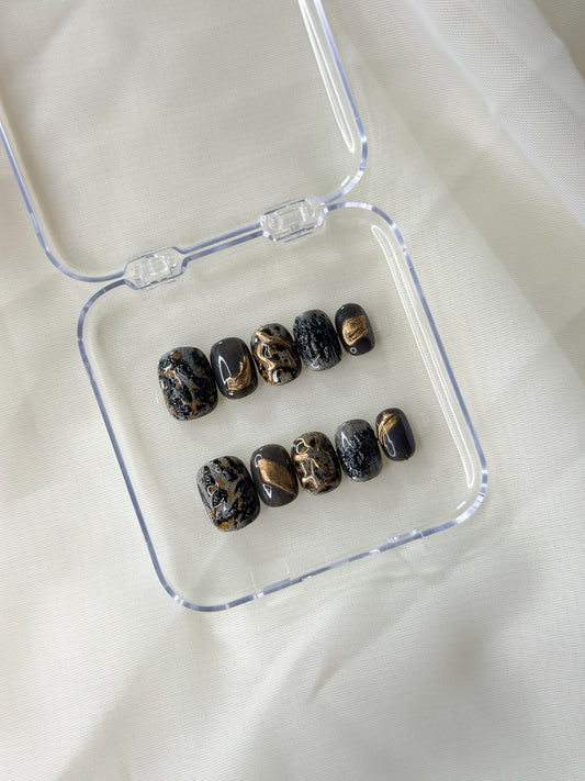 A set of ten decorative press-on nails with a black and gold leopard marbled pattern, displayed inside an open clear plastic case.