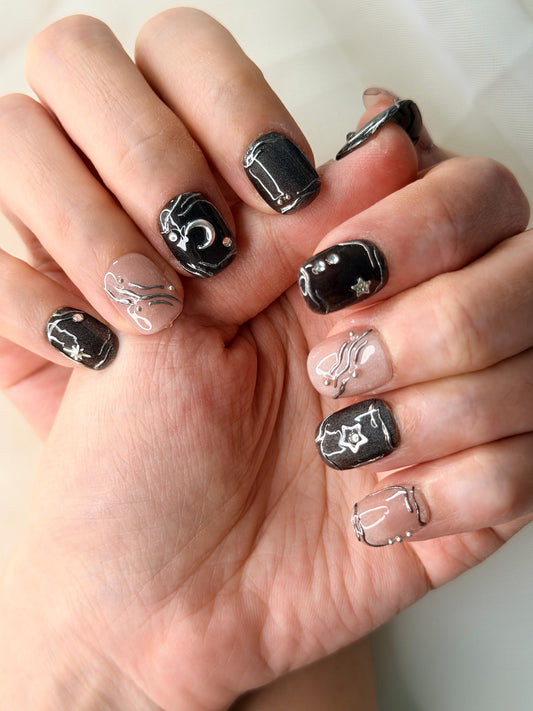 A close-up of a hand showcasing a gothic celestial nail art design. The nails feature a mix of glossy black with silver moon and star illustrations, and sheer nude-pink nails with silver wavy line details.