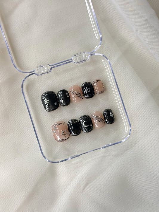 A set of press-on nails displayed in a clear plastic case on a soft white fabric background. The nails feature a mix of glossy black with white celestial designs and a sheer nude pink with black line-art illustrations of stars and moons.