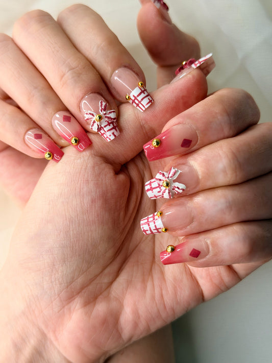 A close-up of hands showcasing a festive Christmas-themed press-on nail set. The nails feature a nude base with red-and-white plaid patterns, small red diamond accents, and miniature white gift boxes with red plaid bows and tiny gold bead details.