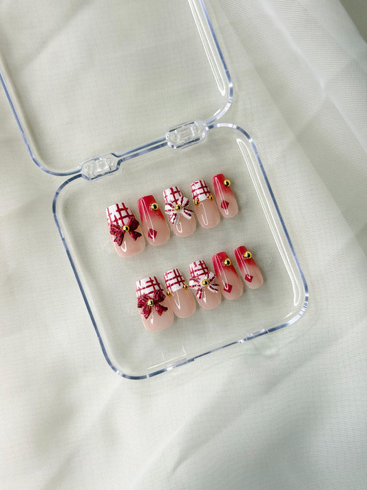 A set of decorative press-on nails displayed inside a clear plastic case on a soft white fabric background. The nails have a pale nude base with red and white plaid patterns, red bows, and small yellow flower accents.