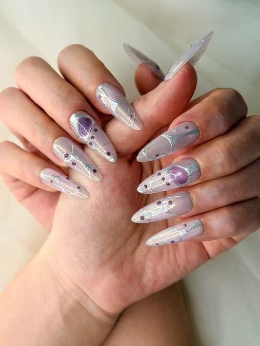 A close-up of hands showcasing long oval, sharp press-on nails with a light lavender and silver glitter base, decorated with sliver geometric lines, small purple dots, and a larger purple heart design on one nail.