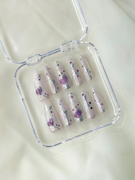 A set of ten press-on nails with a glossy pearl base, decorated with purple droplets, sliver line displayed inside a clear plastic case.