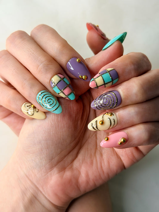 A close-up of a hand featuring colorful, intricate nail art. The manicure features a variety of patterns, including plaid, swirls, stripes, and abstract shapes in pastel shades of purple, pink, teal, and yellow, accented with small gold glitter dots.