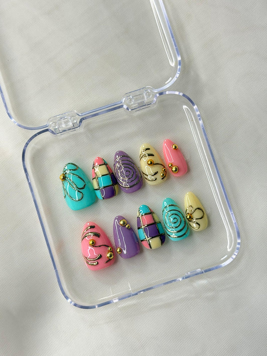 A set of colorful press-on nails displayed inside a clear plastic case features pastel pink, turquoise, purple, and pale yellow colors with metallic gold glitter swirls and small gold bead details.