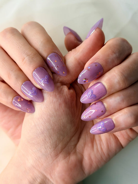 A close-up of hands showcasing a lavender purple press on nails with glittery heart details and subtle flower designs on the nails.
