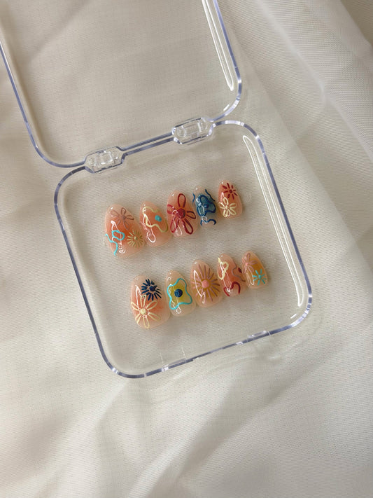 A set of 10 decorative press-on nails displayed inside a clear plastic case on a white fabric background. The nails feature a peachy nude base with colorful floral designs in orange, blue, yellow, and red.