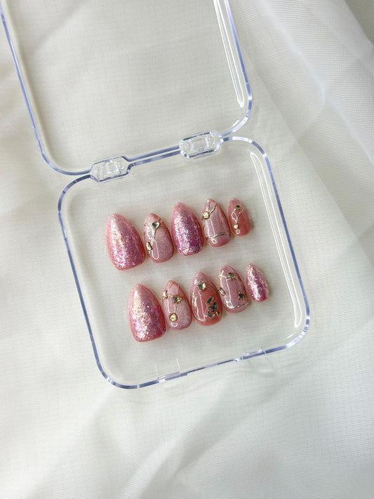 A set of glittery pink press-on nails with small gold and green floral accents, displayed in a clear plastic case.