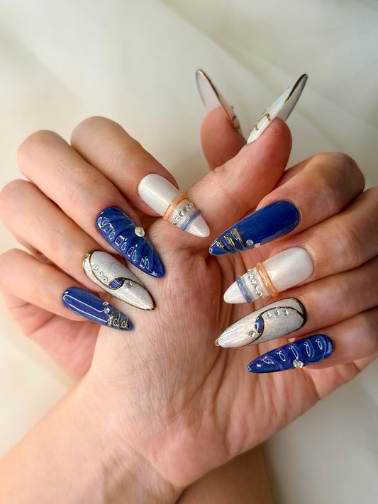 A close-up of hands showcasing a set of long oval-shaped press-on nails with a blue and white celestial design, decorated with metallic gold glitter and small rhinestone accents.