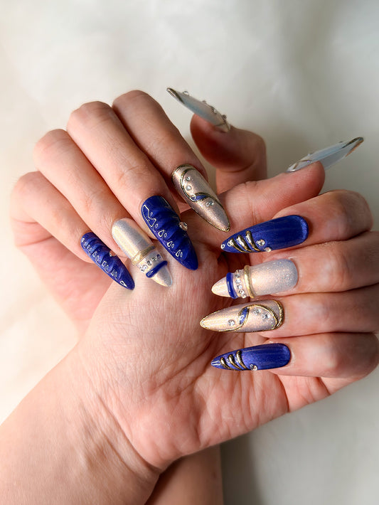 A close-up of hands showcasing a set of long, oval-shaped press-on nails with a blue-and-white celestial design, adorned with metallic gold glitter and small rhinestone accents.