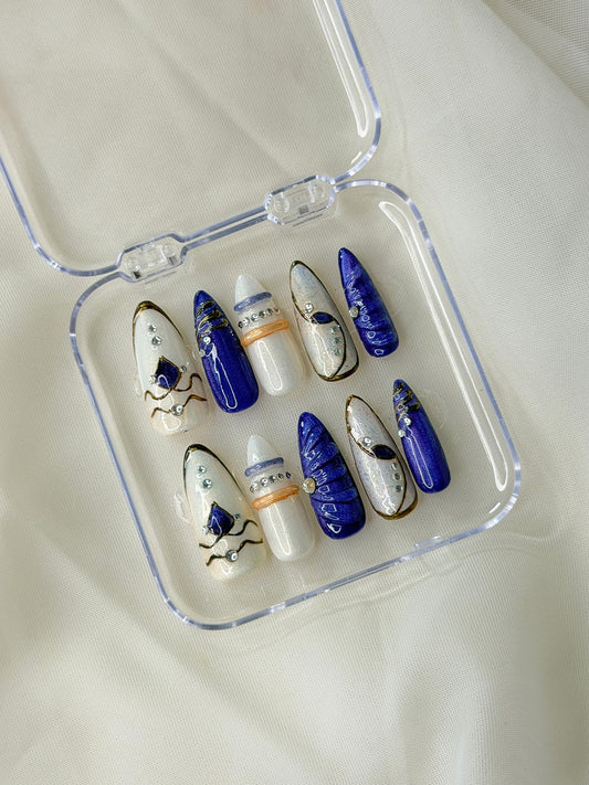 A set of decorative press-on nails displayed in a clear plastic case. The nails feature a mix of deep blue, white, and gold designs with celestial and abstract motifs.