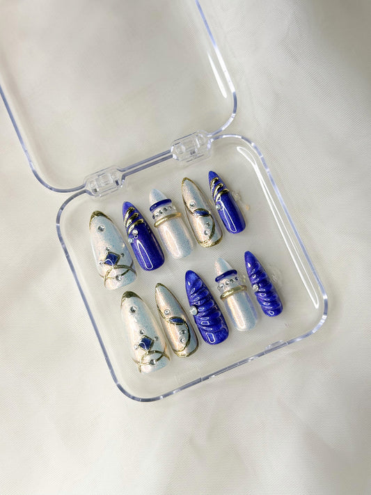 A set of decorative press-on nails displayed in a clear plastic case. The nails feature a mix of deep blue, white, and gold designs with celestial and abstract motifs.