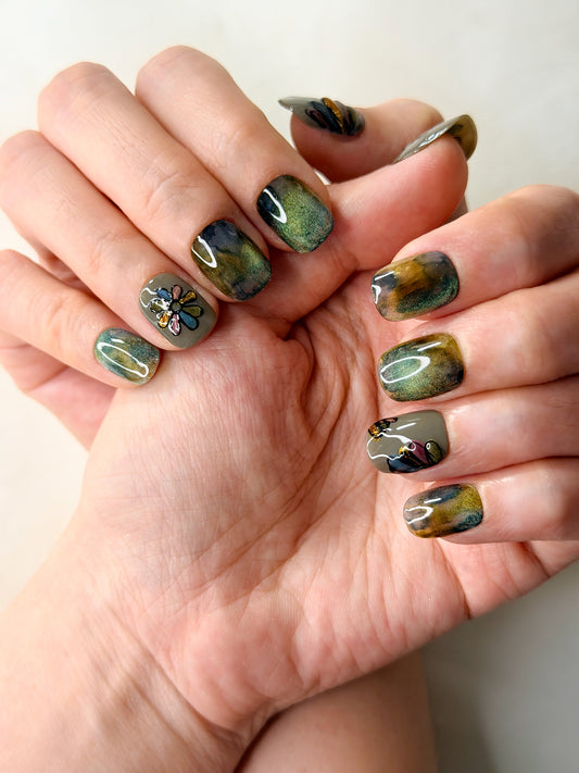 Close-up of hands showcasing a glossy nail art design with a marbled green, brown, and gold pattern and one small floral accent.
