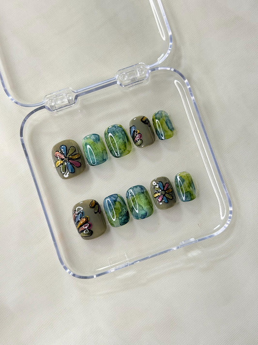 A set of art glass press-on nails displayed in a clear plastic case features a mix of olive green, teal blue, and yellow-green marbled patterns, with some decorated with colorful floral designs.