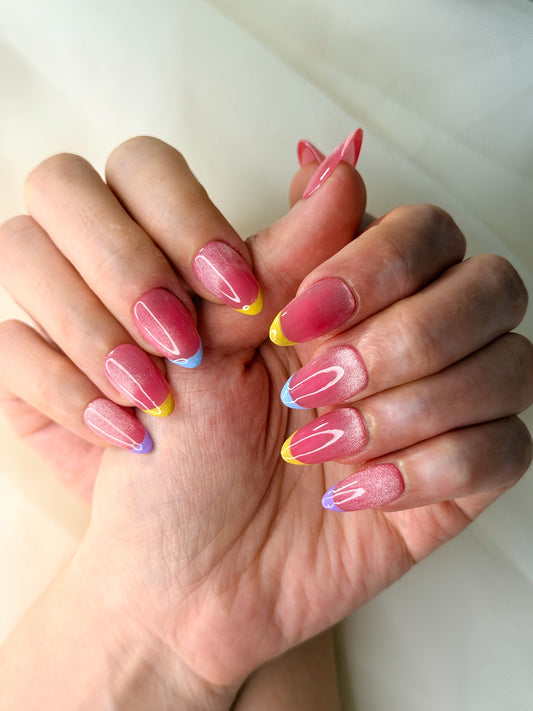 A close-up of hands showcasing short almond press-on nails with a pink glitter base and small colorful french tips accents.