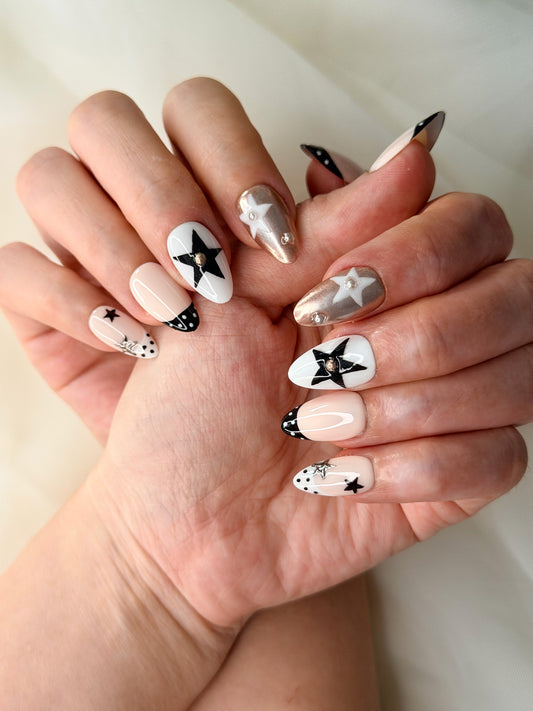 A close-up of hands showcasing a short almond nail art design with a celestial theme. The nails feature white backgrounds with black star details, some with black polka dots, a few with metallic sliver star designs.