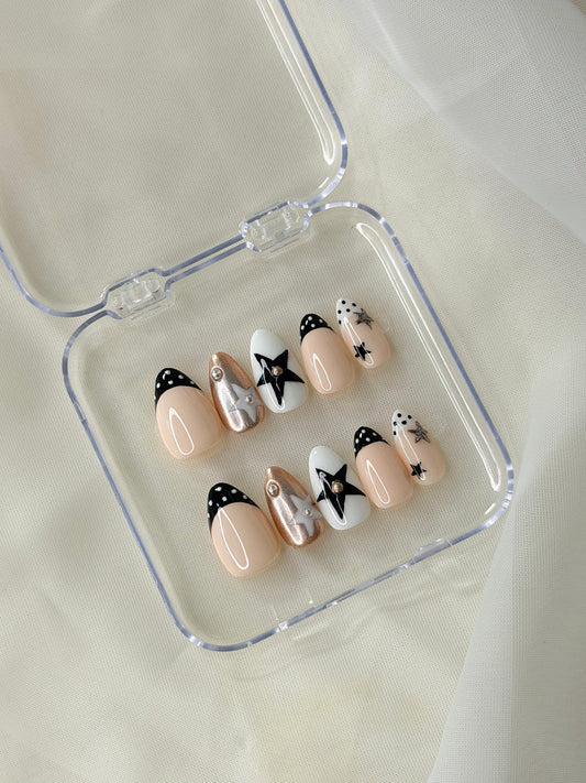 A set of 10 press-on nails features a glossy nude-peach base with black, white, and silver star designs displayed in a clear plastic case.