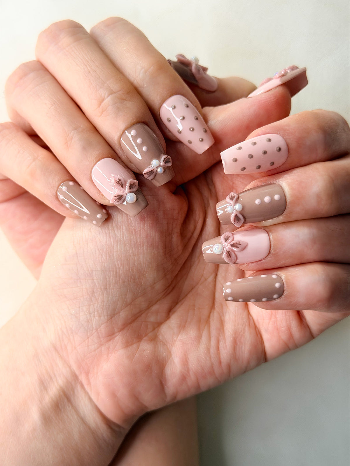 A close-up of hands showcasing coffin-shaped nail set painted in soft pink and taupe brown, decorated with delicate bows, polka dots, and small white pearl accents.
