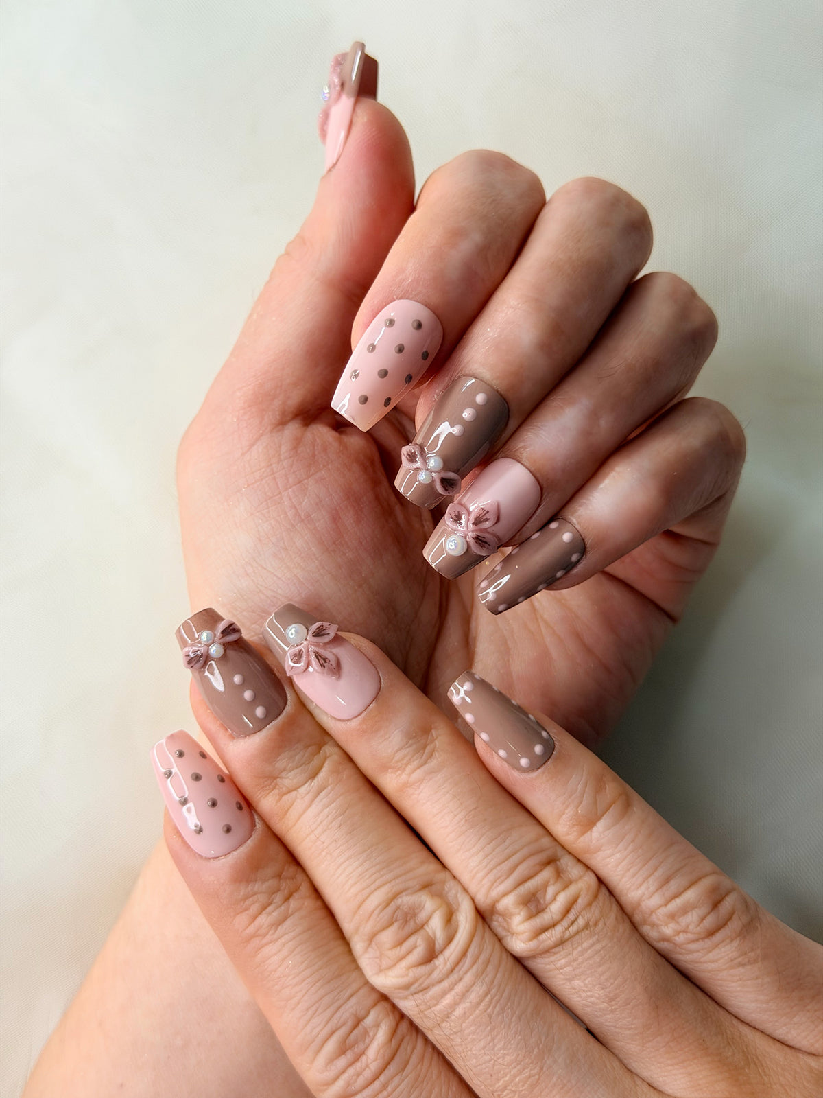 A close-up of a hand with nails featuring a pastel pink and taupe-brown design, decorated with small polka dots, bow details, and white pearls.