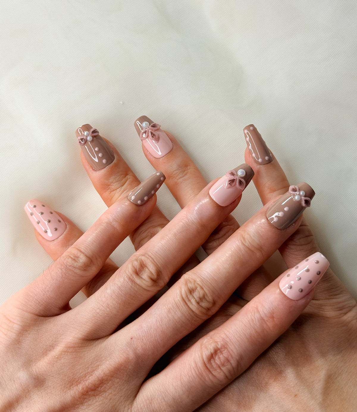 A close-up of a hand showcasing a modern coffin-shaped nail art design in soft pink and taupe brown tones with delicate floral bows, polka dots, and white pearl accents.