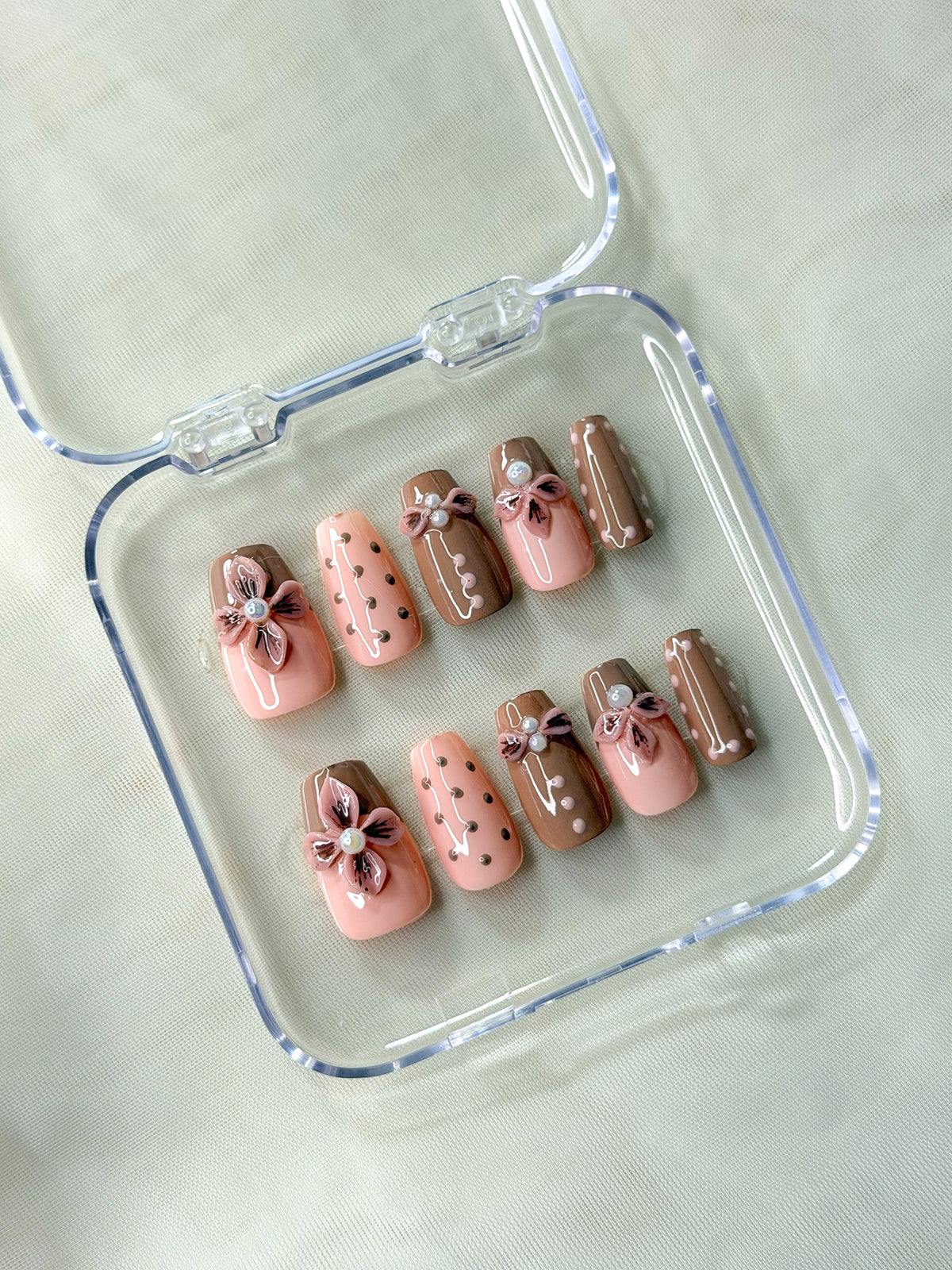 A set of 10 press-on nails is displayed inside a clear plastic case and features a mix of light pink, medium brown, and dark brown colors with decorative floral and polka-dot designs.