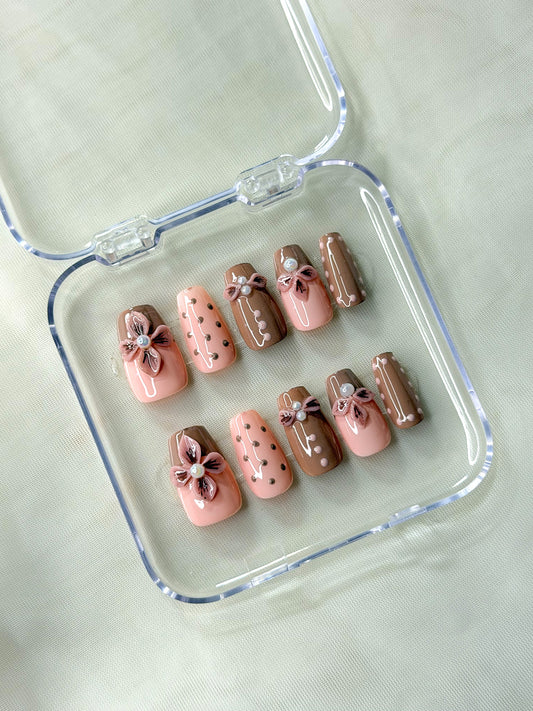 A set of 10 press-on nails is displayed inside a clear plastic case and features a mix of light pink, medium brown, and dark brown colors with decorative floral and polka-dot designs.
